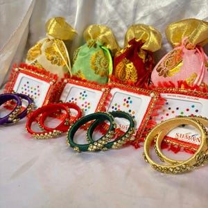 May include: An assortment of colorful bangles and small gift bags. The bangles are in various colors, including purple, red, green, and gold, and are adorned with decorative elements. The gift bags are in gold, green, red, and pink, with gold drawstring closures.