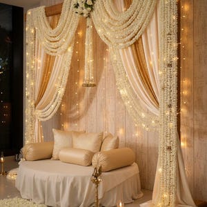 May include: Elegant wedding or event decor featuring a round seating area with cream-colored cushions and a draped skirt. The backdrop includes cream and gold curtains, floral garlands, and string lights, creating a warm, inviting atmosphere.