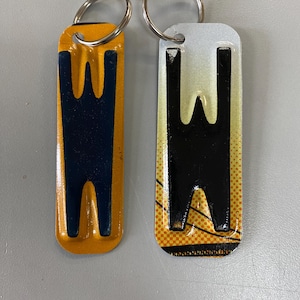K&ouml;nnte beinhalten: Zwei Schl&uuml;sselanh&auml;nger aus Metall mit dem Buchstaben "W" darauf gedruckt. Ein Schl&uuml;sselanh&auml;nger ist gelb mit einem schwarzen "W" und der andere ist wei&szlig; mit einem schwarzen "W".