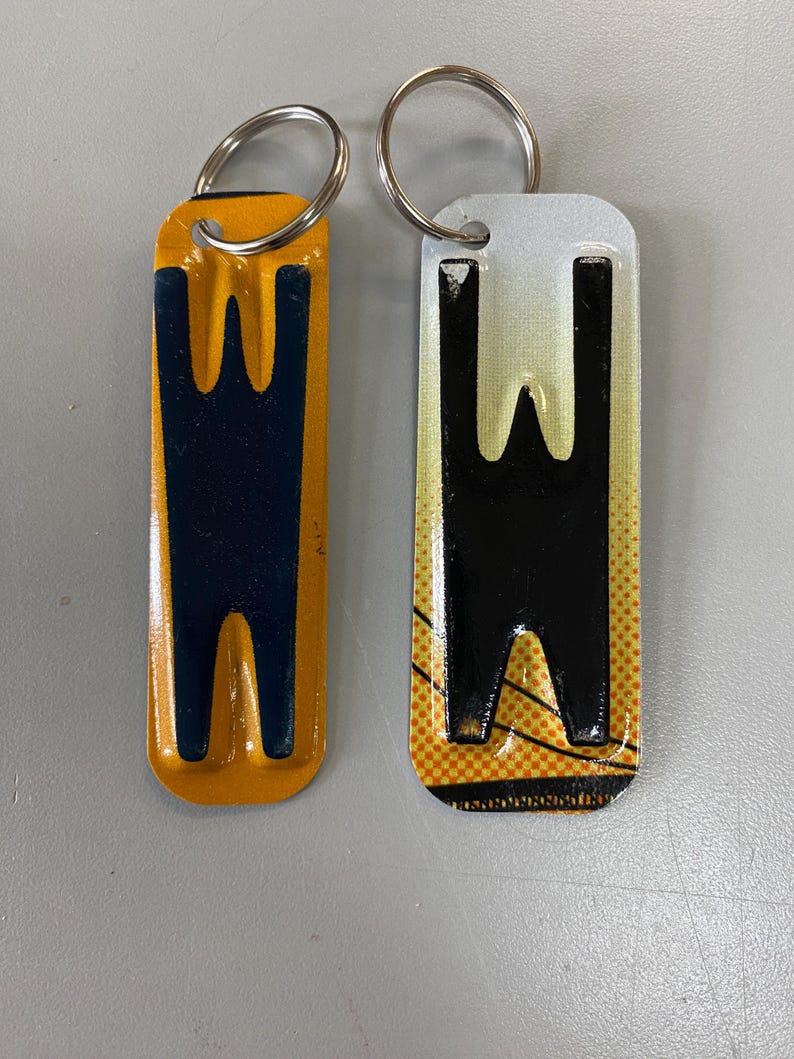 K&ouml;nnte beinhalten: Zwei Schl&uuml;sselanh&auml;nger aus Metall mit dem Buchstaben "W" darauf gedruckt. Ein Schl&uuml;sselanh&auml;nger ist gelb mit einem schwarzen "W" und der andere ist wei&szlig; mit einem schwarzen "W".