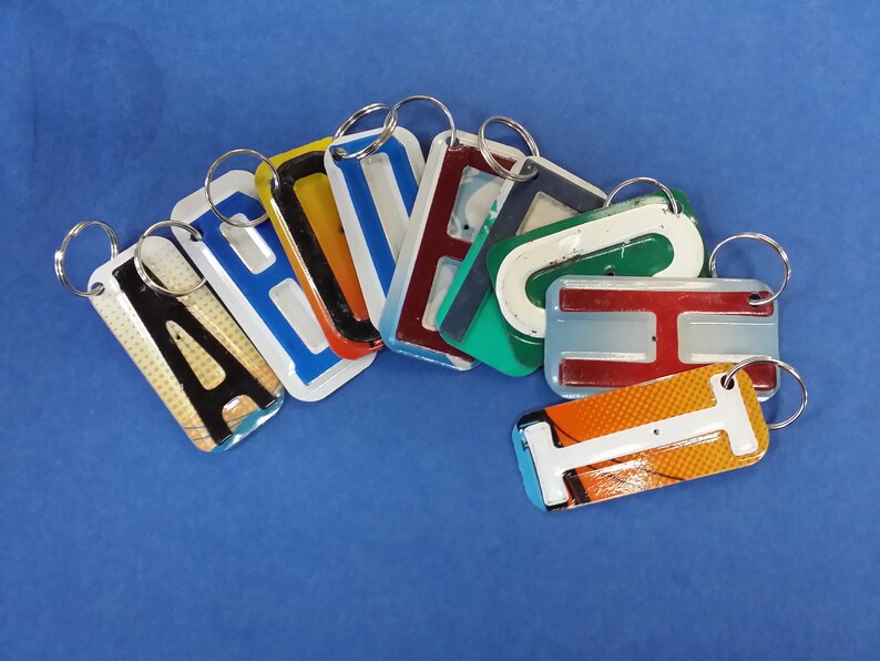 May include: A set of eight keychains, each featuring a different letter of the alphabet. The letters are made of metal and painted in various colors, including red, blue, green, yellow, and orange. The keychains are arranged in a semi-circle on a blue background.