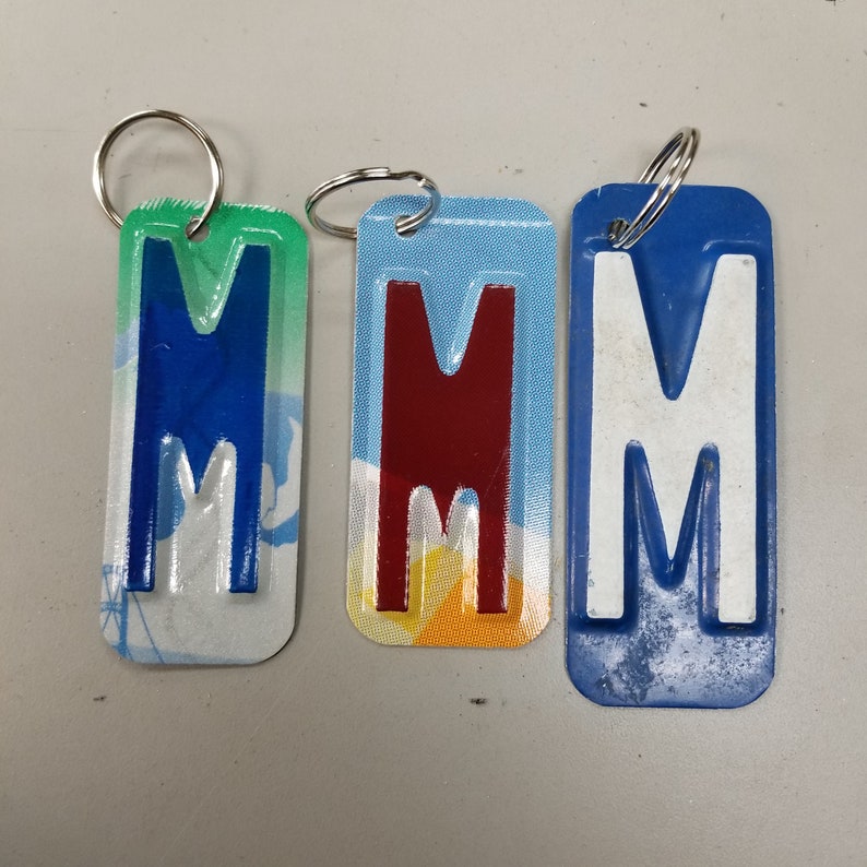 K&ouml;nnte beinhalten: Drei Schl&uuml;sselanh&auml;nger mit dem Buchstaben "M" in verschiedenen Farben. Der erste Schl&uuml;sselanh&auml;nger ist blau mit wei&szlig;em Hintergrund, der zweite ist rot mit gelbem Hintergrund und der dritte ist wei&szlig; mit blauem Hintergrund.