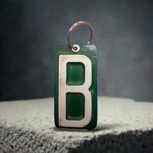 K&ouml;nnte beinhalten: Ein gr&uuml;ner Schl&uuml;sselanh&auml;nger aus Metall mit einem wei&szlig;en Buchstaben B darauf. Der Schl&uuml;sselanh&auml;nger hat einen silbernen Ring.