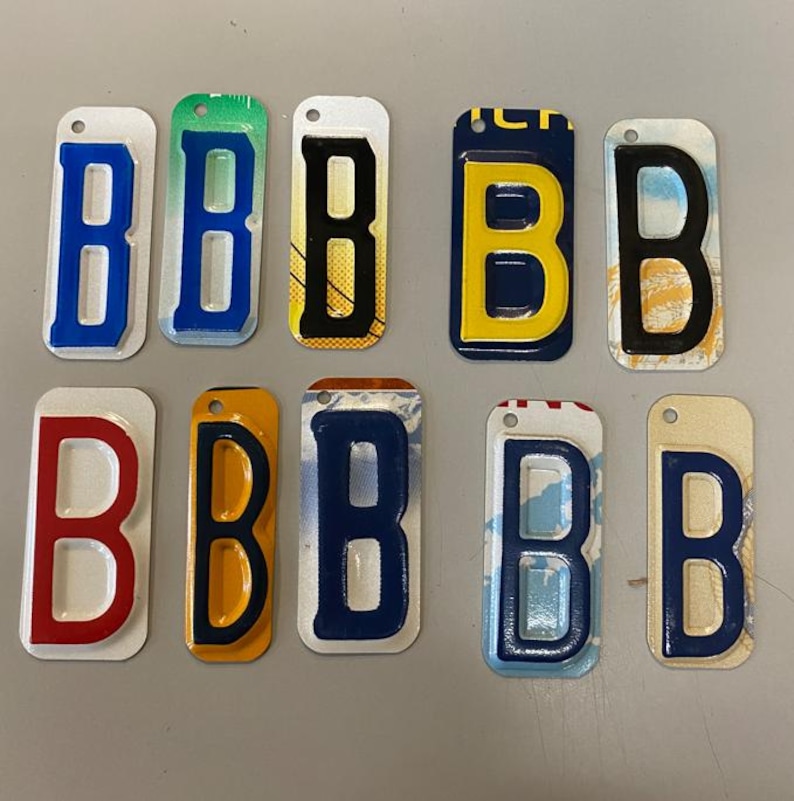 K&ouml;nnte beinhalten: Zehn Schl&uuml;sselanh&auml;nger aus Metall in Form von Nummernschildern, jeweils mit dem Buchstaben "B" in einer anderen Farbe und einem anderen Design. Die Schl&uuml;sselanh&auml;nger sind in zwei Reihen zu f&uuml;nf angeordnet. Die Farben umfassen Rot, Blau, Gelb, Schwarz und Wei&szlig;.