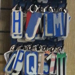 K&ouml;nnte beinhalten: Ein Satz von 26 Schl&uuml;sselanh&auml;ngern mit Buchstaben des Alphabets, jeder Buchstabe ist auf wei&szlig;em Hintergrund mit blauem Rand. Die Buchstaben sind in alphabetischer Reihenfolge angeordnet und h&auml;ngen an einem Metallgestell.