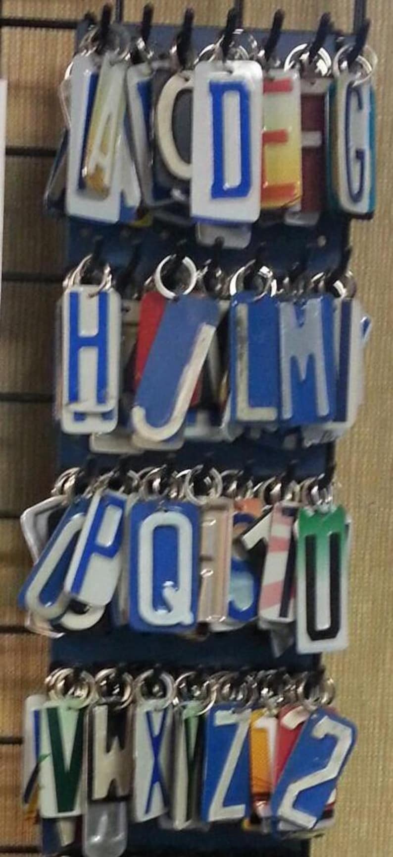K&ouml;nnte beinhalten: Ein Satz von 26 Schl&uuml;sselanh&auml;ngern mit Buchstaben des Alphabets, jeder Buchstabe ist auf wei&szlig;em Hintergrund mit blauem Rand. Die Buchstaben sind in alphabetischer Reihenfolge angeordnet und h&auml;ngen an einem Metallgestell.