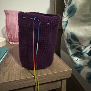 May include: A handmade, dark purple crochet basket with colorful ribbon drawstring. The basket is cylindrical and sits on a wooden nightstand. A pink glass vase is visible in the background.