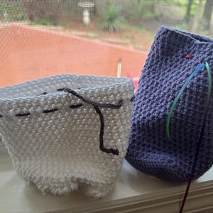 May include: Two handmade crochet containers. One is white with dark gray stitching and a drawstring. The other is purple with a drawstring and colorful ribbons. Both are sitting on a windowsill.