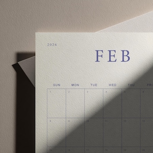 May include: A close-up of a calendar page for February 2026. The word "FEB" is in a dark blue font. The days of the week are listed across the top, with numbered boxes below. The calendar is on a textured, off-white paper.