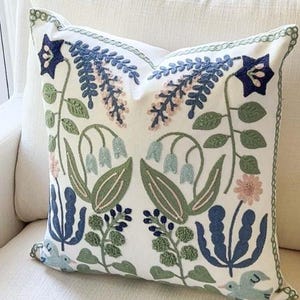 Könnte beinhalten: Quadratisches weißes Kissen mit gesticktem Blumendesign. Das Kissen hat blaue, grüne und rosa Blumen, Blätter und Stiele. Das Kissen hat eine grün genähte Bordüre und liegt auf einem weißen Stuhl.