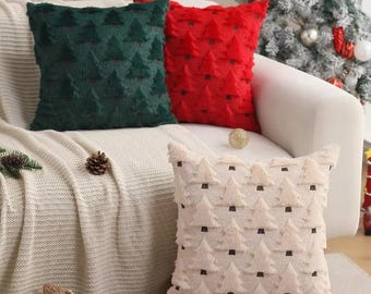 May include: Three decorative cushions with a Christmas tree pattern. One is dark green, one is red, and the other is off-white. The cushions are on a white sofa with a knitted blanket. A Christmas tree is in the background.