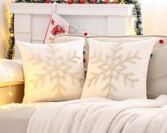 Può includere: Due cuscini decorativi bianchi con ricami a forma di fiocco di neve. I cuscini sono su un divano color crema con una soffice coperta bianca. Lo sfondo include un caminetto decorato per Natale.