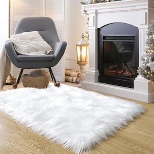 May include: A fluffy, rectangular white faux fur rug on a hardwood floor. The rug is in a living room setting, with a gray armchair, a fireplace, and a Christmas tree in the background. The rug adds a cozy and warm touch to the room.