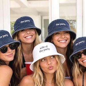 Könnte beinhalten: Eine Gruppe von Frauen trägt Jeans-Eimerhüte mit dem gestickten Text "the party". Eine Frau trägt einen weißen Hut mit der Aufschrift "wife of the party". Die Frauen lächeln und tragen Sonnenbrillen.