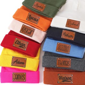 Op de afbeelding: Een collectie van twaalf gebreide mutsen in diverse kleuren zoals olijfgroen, oranje, roze, rood, geel, felroze, wit, zwart, lichtblauw, grijs en bruin, elk voorzien van een bruine leren patch met een unieke naam erin gegraveerd.