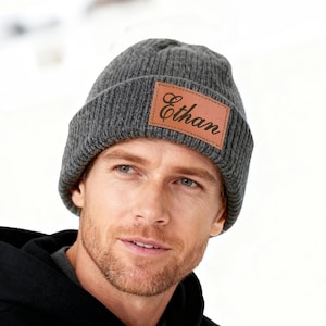 May include: Gray knit beanie with a brown leather patch. The patch is rectangular and features the name "Ethan" in a script font. The beanie is a ribbed knit style.