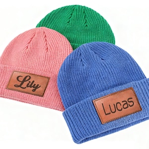 May include: Three knit beanies in pink, green, and blue. Each beanie has a brown leather patch with a name: "Lily" on the pink beanie and "Lucas" on the blue beanie. The green beanie is plain.