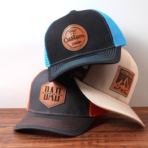 May include: Three baseball caps with leather patches. One black cap has a blue mesh back and a patch that says "Your Custom Design". Another black cap has an orange mesh back and a patch that says "DAD". The third cap is tan with a mountain patch.