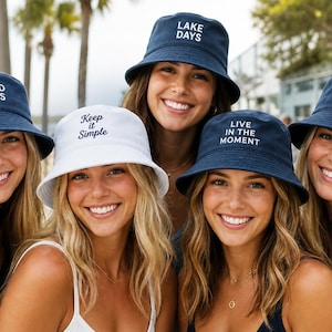 K&ouml;nnte beinhalten: Eine Auswahl von f&uuml;nf Fischerh&uuml;ten in Marineblau und Wei&szlig;. Die marineblauen H&uuml;te haben wei&szlig;e Stickereien mit den Texten GOOD VIBES, LAKE DAYS, LIVE IN THE MOMENT und GOOD TIMES mit einem Smiley. Der wei&szlig;e Hut tr&auml;gt den Text Keep it Simple.