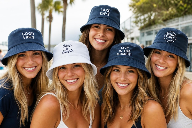 K&ouml;nnte beinhalten: Eine Auswahl von f&uuml;nf Fischerh&uuml;ten in Marineblau und Wei&szlig;. Die marineblauen H&uuml;te haben wei&szlig;e Stickereien mit den Texten GOOD VIBES, LAKE DAYS, LIVE IN THE MOMENT und GOOD TIMES mit einem Smiley. Der wei&szlig;e Hut tr&auml;gt den Text Keep it Simple.
