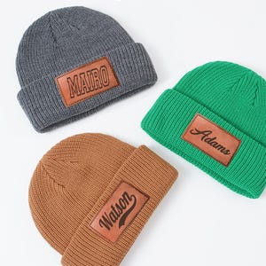 May include: Three knit beanies in gray, green, and brown. Each beanie has a rectangular leather patch with a name: "MAIRO", "Adams", and "Watson". The beanies are displayed on a white surface.