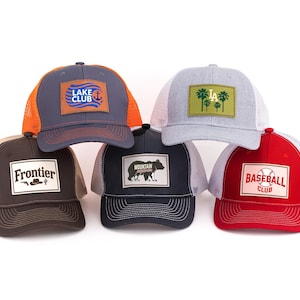 Könnte beinhalten: Eine Sammlung von Trucker-Caps mit verschiedenen Designs. Die Caps haben Mesh-Rücken und Stofffronten mit Aufnähern. Designs sind "Lake Club", "LA", "Frontier", ein Bär und "Baseball Club". Farben sind Grau, Orange, Braun, Schwarz und Rot.