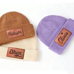 Può includere: Tre berretti lavorati a maglia in marrone, crema e viola. Ogni berretto ha una toppa rettangolare in pelle con un nome: "Madison", "Ethan" e "Olivia". La toppa "Olivia" include un simbolo a cuore. I berretti sono ripiegati sull'orlo.
