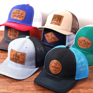Può includere: Una collezione di cappellini da camionista in vari colori, tra cui blu, rosso, nero e verde. Ogni cappellino presenta una toppa in pelle con testo o un logo. I cappellini hanno schienali in rete e visiere curve, con frasi come "The World is Yours" e "Best Dad".