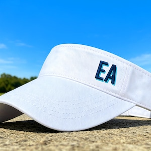 May include: White visor with the embroidered letters "EA" in blue. The visor is made of a smooth fabric and has a curved brim. The background features a blue sky and green foliage.