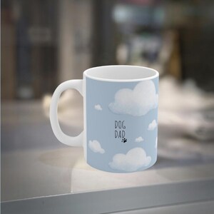 Peut inclure: Une tasse en céramique blanche avec un fond bleu clair et des illustrations de nuages blancs. La tasse porte l'inscription "DOG DAD" avec une empreinte de patte. La tasse a une anse blanche.