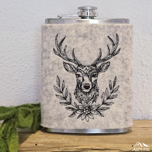 Nahaufnahme eines Flachmanns in grauer Filzhülle mit einem gestickten Hirschkopf-Porträt im Lorbeerkranz.

Close-up of a flask in a grey felt sleeve featuring an embroidered stag head portrait in a wreath.