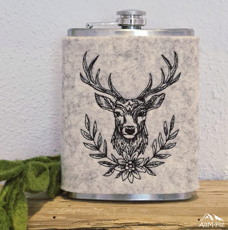 Nahaufnahme eines Flachmanns in grauer Filzhülle mit einem gestickten Hirschkopf-Porträt im Lorbeerkranz.

Close-up of a flask in a grey felt sleeve featuring an embroidered stag head portrait in a wreath.