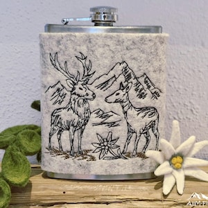 Flachmann in grauer Filzhülle mit einer gestickten Szene von Hirsch und Reh vor Bergen und Edelweiß.

Flask in a grey felt sleeve with an embroidered scene of a stag and deer in front of mountains.