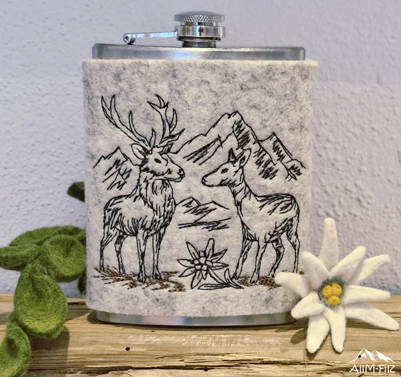Flachmann in grauer Filzhülle mit einer gestickten Szene von Hirsch und Reh vor Bergen und Edelweiß.

Flask in a grey felt sleeve with an embroidered scene of a stag and deer in front of mountains.