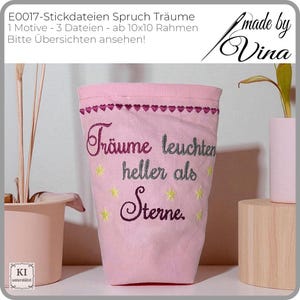 Könnte beinhalten: Ein rosa Stoffbehälter mit gesticktem Text "Träume leuchten heller als Sterne" und Sternen. Der obere Rand hat eine Reihe von herzförmigen Stickereien. Der Behälter wird mit anderen dekorativen Gegenständen präsentiert.