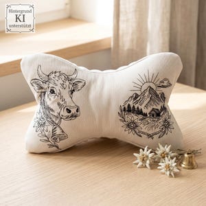 Knochenförmiges Nackenkissen mit gesticktem Kuh-Porträt und Berghütten-Motiv auf einem Holztisch.

Bone-shaped neck pillow with embroidered cow portrait and mountain cabin design on a wooden table.
