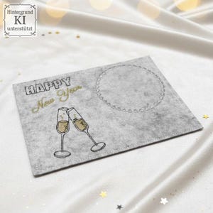Puede incluir: Tarjeta de fieltro gris con bordado negro y dorado "HAPPY New Year". Dos copas de champ&aacute;n brindando. Un c&iacute;rculo delineado a la derecha. Fondo de sat&eacute;n blanco. Ideal para felicitaciones de A&ntilde;o Nuevo.