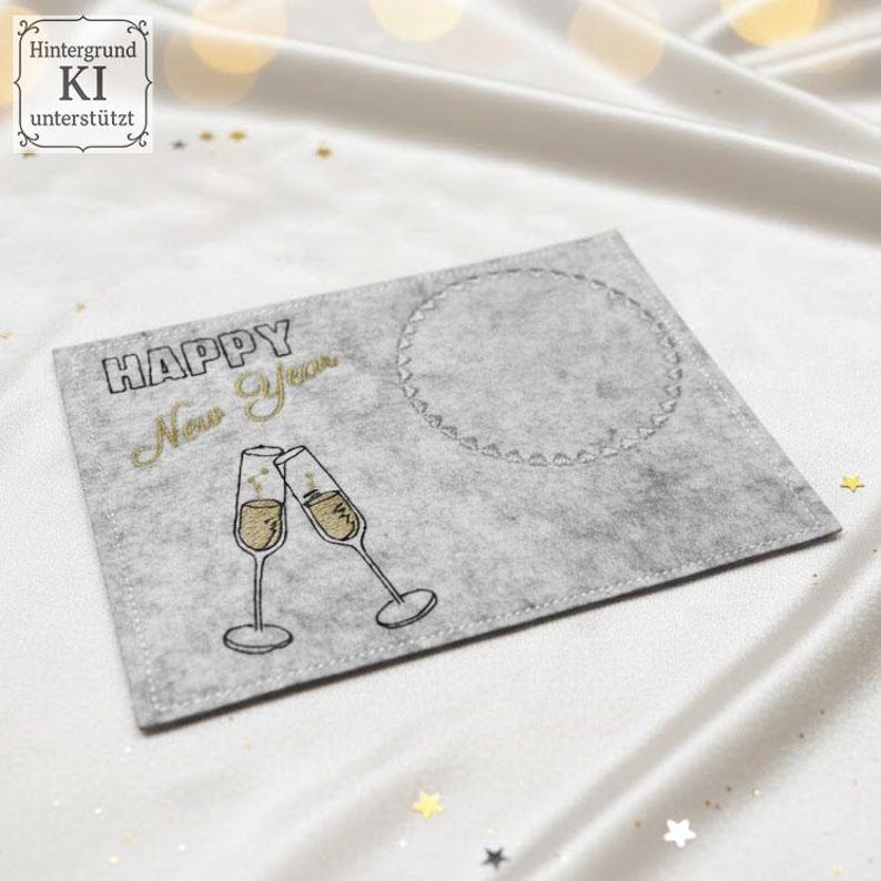 Puede incluir: Tarjeta de fieltro gris con bordado negro y dorado "HAPPY New Year". Dos copas de champ&aacute;n brindando. Un c&iacute;rculo delineado a la derecha. Fondo de sat&eacute;n blanco. Ideal para felicitaciones de A&ntilde;o Nuevo.