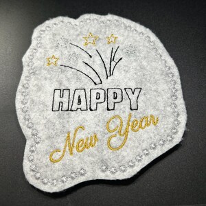 Puede incluir: Decoraci&oacute;n de A&ntilde;o Nuevo de fieltro gris con texto y dise&ntilde;o bordados. Las palabras "HAPPY New Year" est&aacute;n cosidas con hilo negro y dorado. El dise&ntilde;o incluye fuegos artificiales y estrellas.
