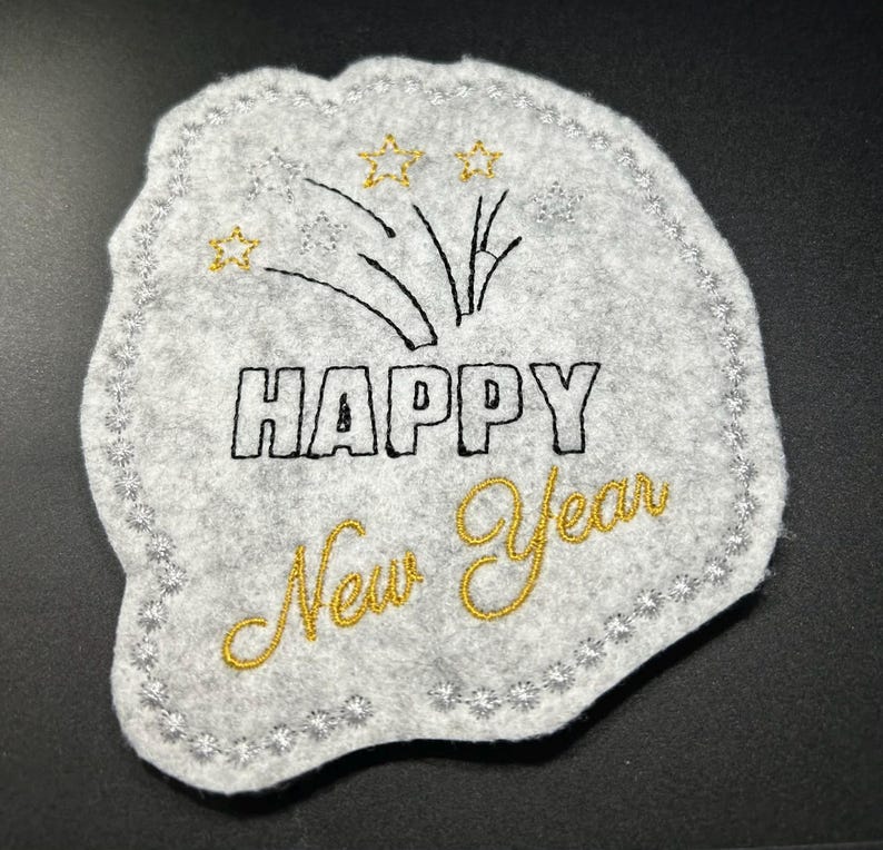 Puede incluir: Decoraci&oacute;n de A&ntilde;o Nuevo de fieltro gris con texto y dise&ntilde;o bordados. Las palabras "HAPPY New Year" est&aacute;n cosidas con hilo negro y dorado. El dise&ntilde;o incluye fuegos artificiales y estrellas.
