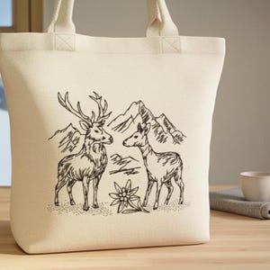 Helle Canvas-Einkaufstasche mit gesticktem Hirsch- und Reh-Motiv vor Bergsilhouette auf einem Holztisch.

Light canvas tote bag with an embroidered stag and deer motif in front of a mountain silhouette.