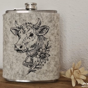 Flachmann in grauer Filzhülle mit gesticktem Kuhkopf-Porträt mit Glocke und Edelweißblüte.

Flask in a grey felt sleeve with an embroidered cow portrait wearing a bell and an edelweiss flower.