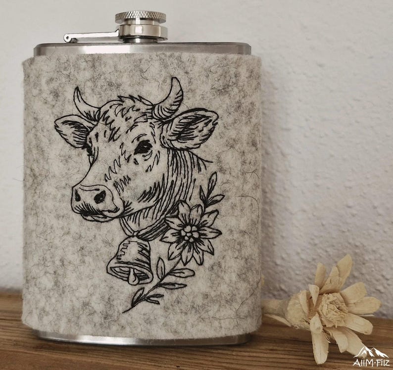 Flachmann in grauer Filzhülle mit gesticktem Kuhkopf-Porträt mit Glocke und Edelweißblüte.

Flask in a grey felt sleeve with an embroidered cow portrait wearing a bell and an edelweiss flower.