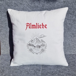 Weißes Kissen mit dem roten Schriftzug Almliebe und einer filigranen Stickerei einer Berghütte im Scribble-Stil.

White throw pillow with red embroidered text Almliebe and a delicate mountain cabin design in scribble style.