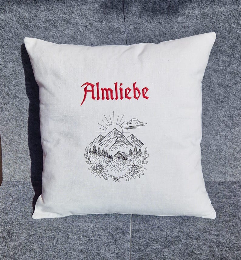 Weißes Kissen mit dem roten Schriftzug Almliebe und einer filigranen Stickerei einer Berghütte im Scribble-Stil.

White throw pillow with red embroidered text Almliebe and a delicate mountain cabin design in scribble style.