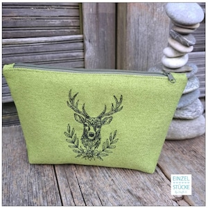 Hellgrünes Kosmetiktäschchen aus Webstoff mit einem gestickten Hirschkopf vor einer Steinpyramide.

Light green woven cosmetic bag with an embroidered stag head next to a stack of balanced stones.