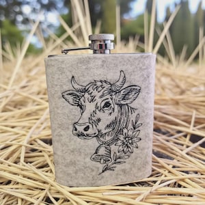 Flachmann in grauer Filzhülle mit Kuh-Stickerei auf einem Untergrund aus trockenem Stroh.

Flask in a grey felt sleeve with cow embroidery placed on a bed of dry straw.