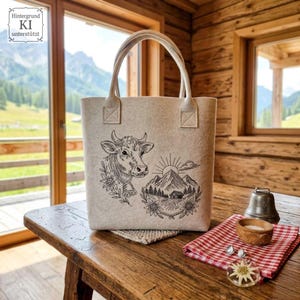 Große Filz-Tragetasche mit Kuh- und Berghütten-Stickerei in einer rustikalen Holzhütte dekoriert.

Large felt tote bag with cow and mountain cabin embroidery displayed inside a rustic wooden cabin.
