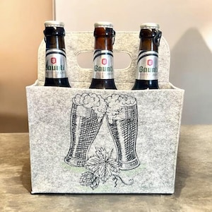 Filzträger für Bierflaschen mit gestickten vollen Biergläsern und Hopfenblättern vor hellem Hintergrund.

Felt beer bottle carrier with embroidered full beer glasses and hop leaves on a light background.