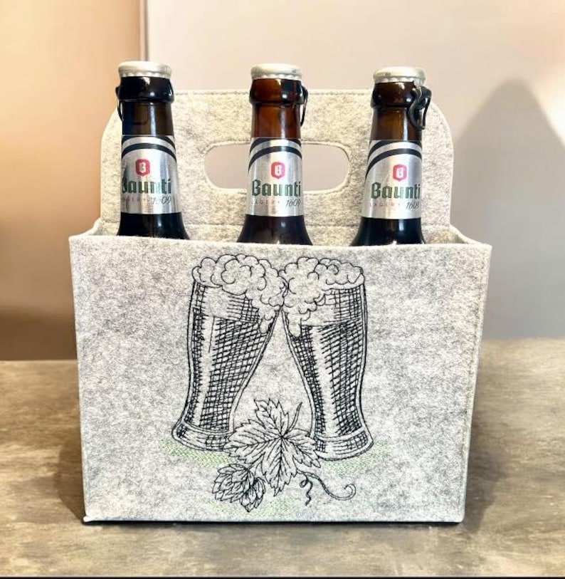 Filzträger für Bierflaschen mit gestickten vollen Biergläsern und Hopfenblättern vor hellem Hintergrund.

Felt beer bottle carrier with embroidered full beer glasses and hop leaves on a light background.